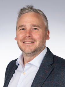 A man with short, grey hair in a dark suit jacket and white shirt.