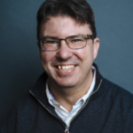 A headshot of Neil Campbell, a man wearing a white collar with a blue zipped sweatshirt. He has short, dark hair and black framed, rectangular glasses.
