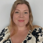 A woman with long, blonde hair worn down, with a v-neck white and black flower blouse in a bold patter. Anna is smiling at the camera.