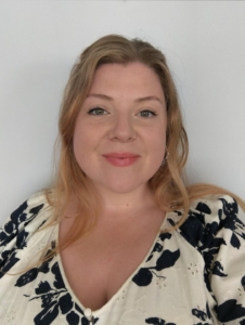 A woman with long, blonde hair worn down, with a v-neck white and black flower blouse in a bold patter. Anna is smiling at the camera.